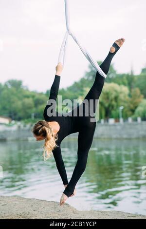 Donna aeralista esegue ginnastica spaccata su seta aerea appesa sullo sfondo di fiume, cielo e alberi. Foto Stock