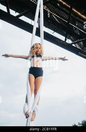 La bambina aerialista esegue acrobazie su seta aerea appesa attaccata al supporto del ponte contro lo sfondo del cielo. Foto Stock