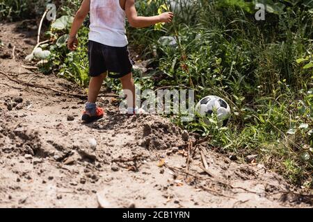 Vista ritagliata del ragazzo afro-americano che si trova vicino al calcio palla in erba vicino a strada sporca in via urbana Foto Stock