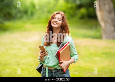 Bella Ginger Girl Student Holding smartphone e libri di lavoro, rilassarsi all'aperto dopo le lezioni Foto Stock