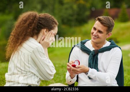 Ritratto di sorridente giovane uomo che apre ring box mentre si propone a fidanzata durante la data romantica all'aperto Foto Stock