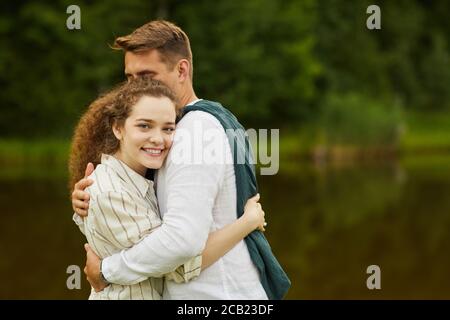 Vita in su ritratto di giovane coppia amorevole abbracciando e guardando la macchina fotografica mentre si sta in piedi vicino al lago all'aperto in estate, copia spazio Foto Stock