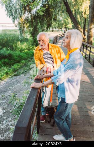 Donna anziana in piedi vicino al marito sorridente con il cane pug sopra guinzaglio sul ponte nel parco Foto Stock