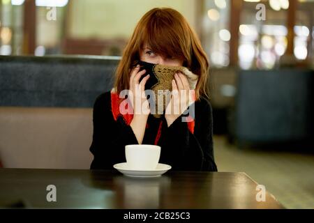 Donna congela, in caffè si riscalda con tazza di bevanda calda, avvolgente, nasconde il viso in sciarpa calda Foto Stock