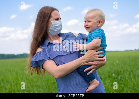 Madre felice in maschera medica protettiva tiene il suo bambino sulle armi in natura in estate. Fine del coronavirus, infanzia e concetto di famiglia. Foto Stock