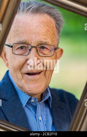 Vista testa e spalle di un pensionato maschile che guarda attraverso una cornice da cartolina tenuta ad angolo davanti a uno sfondo sfocato di un parco estivo. Foto Stock