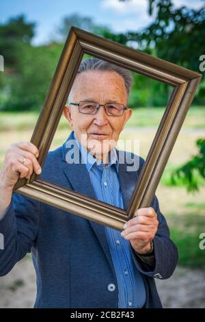 Vista superiore del corpo di un pensionato maschile che guarda attraverso una cornice di foto tenuta ad un angolo di fronte a uno sfondo sfocato di un parco estivo. Foto Stock