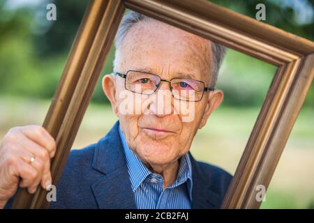 Vista testa e spalle di un pensionato maschile che guarda attraverso una cornice da cartolina tenuta ad angolo davanti a uno sfondo sfocato di un parco estivo. Foto Stock