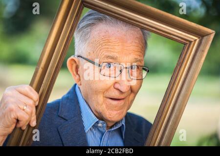 Vista testa e spalle di un pensionato di 80 anni con occhiali dall'aspetto malizioso attraverso una cornice di foto tenuta ad un angolo davanti di un b offuscato Foto Stock