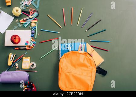 vista dall'alto dello zaino con notebook, matite colorate e forbici vicino alle forniture scolastiche su lavagna verde Foto Stock