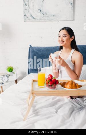 Fuoco selettivo della donna asiatica positiva che tiene la tazza del caffè vicino colazione sul vassoio sul letto Foto Stock