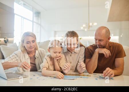 Ritratto dai toni caldi della famiglia moderna con due bambini che risolvono il puzzle insieme mentre si gode il tempo in casa, spazio di copia Foto Stock