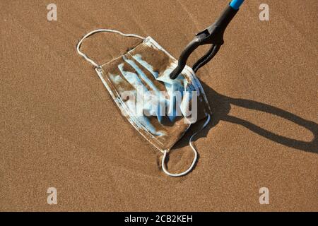 closeup di qualcuno che utilizza un estensione reach per raccogliere un maschera chirurgica blu utilizzata gettata sulla sabbia bagnata di il mare o riportato dall'oceano a th Foto Stock