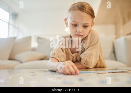 Ritratto dai toni caldi di cute bambina che risolve il puzzle da solo mentre si siede sul divano in un interno domestico minimo, spazio di copia Foto Stock