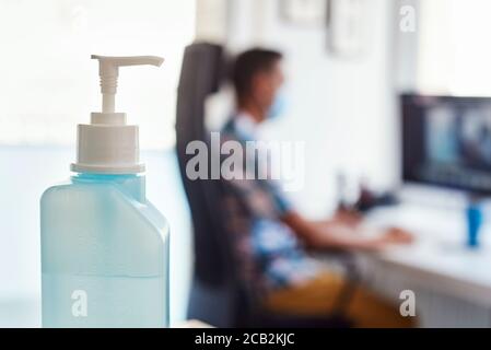 primo piano di una bottiglia di disinfettante per le mani e di un giovane uomo seduto alla scrivania dell'ufficio che indossa una maschera sullo sfondo Foto Stock