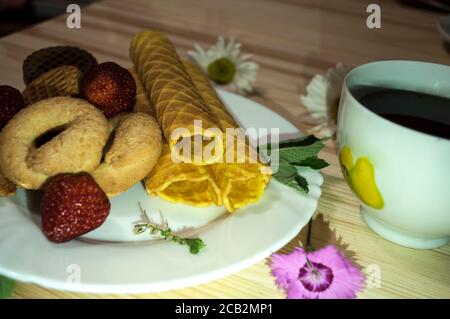 Cialde, fragole, biscotti e una tazza di caffè. Composizione su sfondo di legno chiaro decorata con fiori di garofano e camomilla Foto Stock