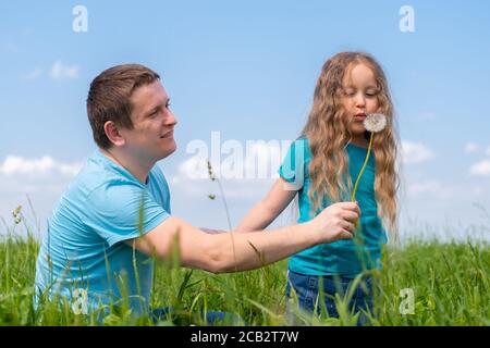 padre e figlie trascorrono del tempo. Papà e figlie che giocano insieme su campo erba verde in estate. Ragazza che soffia su dente di leone Foto Stock