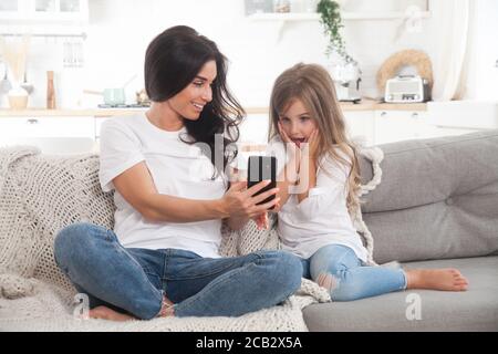 Giovane mamma e bambina che giocano online a casa utilizzando lo smartphone, chattando video o guardando film. Formazione a distanza. Video Foto Stock