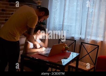 giovane uomo caucasico aiuta suo figlio a studiare, a fare i compiti a casa, utilizzando un computer portatile moderno e libri di esercizi Foto Stock