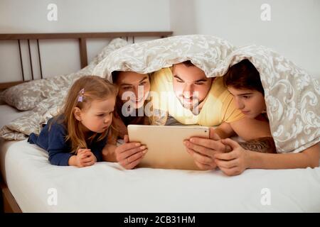 famiglia caucasica, padre madre e bambini che giacciono sotto la coperta a letto guardando video su tablet, tempo libero a casa. Foto Stock