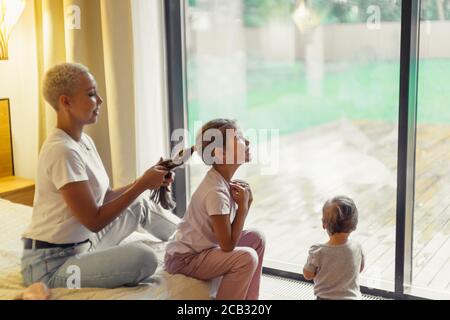 Giovane donna treccia capelli a figlia, seduta a letto nel fine settimana. Finestra panoramica di sfondo, baby stand di fronte alla finestra Foto Stock