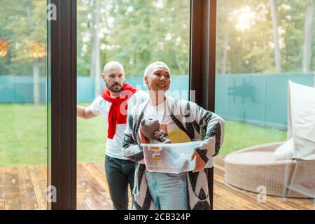 Allegro positivo coppia un casual vestiti che tengono scatole snter nuova casa. Acquisto nuova casa con grande finestra vista sul giardino Foto Stock