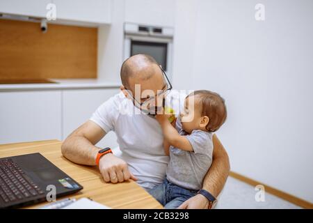 Padre guarda il figlio con tenerezza mentre il bambino tiene la mela verde. Computer portatile sul tavolo. Concetto di famiglia Foto Stock