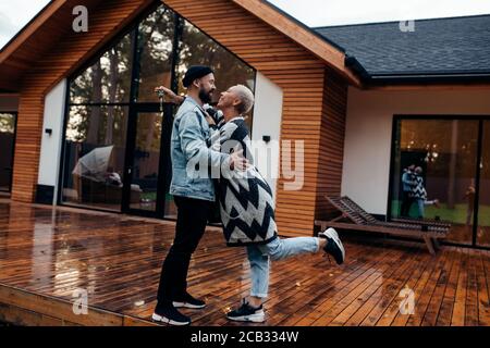 Dolce coppia famiglia baciare stand vicino a nuova casa di legno. Uomo e donna in abiti casual Foto Stock