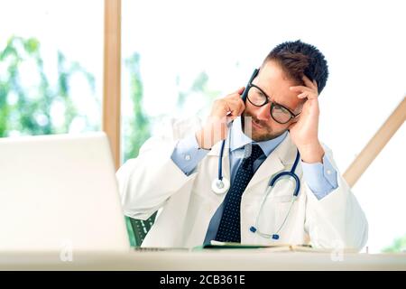 Un colpo di medico maschile stressato seduto alla scrivania dietro il suo laptop e facendo una chiamata nella stanza del medico. Foto Stock