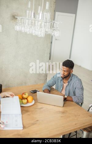 Lavoratore autonomo, giovane uomo afro-americano dalla pelle scura che lavora sul laptop mentre si siede al tavolo in soggiorno Foto Stock