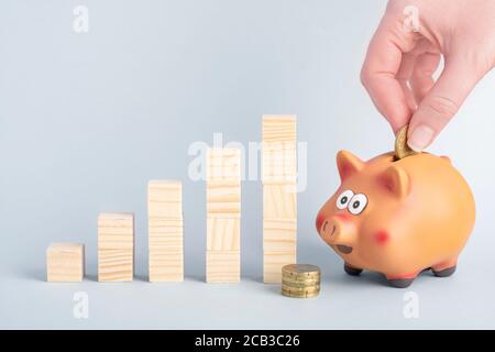 Una mano che puttila una moneta in una banca del piggy e. concetto grafico di crescita da blocchi di legno Foto Stock