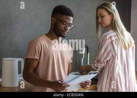 Una coppia mista di progettisti sviluppa un progetto di start-up a casa, tenendo in mano grafici e calcoli su fogli di carta. Famiglia imprenditori coll Foto Stock