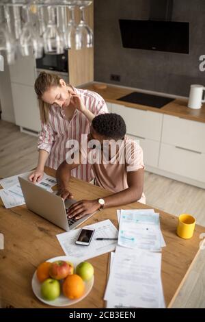 Giovani interracial giovane utilizzando laptop insieme avendo prima colazione a casa, uomo africana ed europea donna bionda alla ricerca di nuove offerte di vendita o shoppi Foto Stock