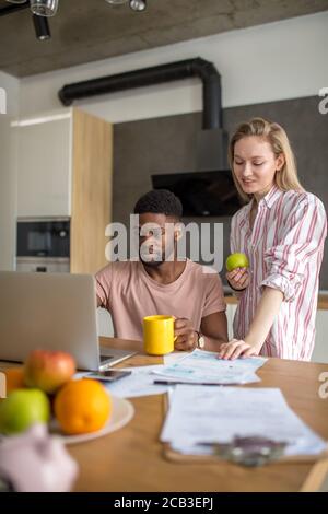 Giovane studente africano termina la sua presentazione in last minute, durante la prima colazione e la sua bionda moglie lo aiuta con consigli, in piedi accanto a Foto Stock