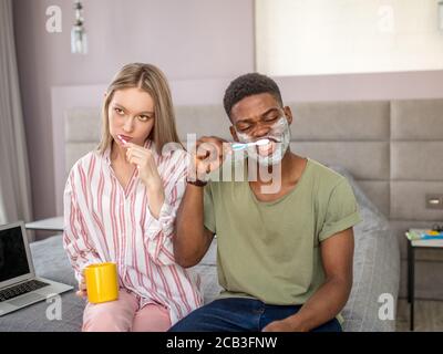 Giovane coppia assonnata nel lavaggio dei denti al mattino, l'uomo sta facendo la crema da barba sulle guance. L'igiene dentale è importante. Foto Stock