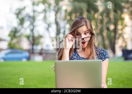 Mi sono hacked. Donna studentesca scioccata con computer portatile che guarda lo schermo che tiene gli occhiali giù in shock seduta nel parco pubblico del campus fuori verde b Foto Stock