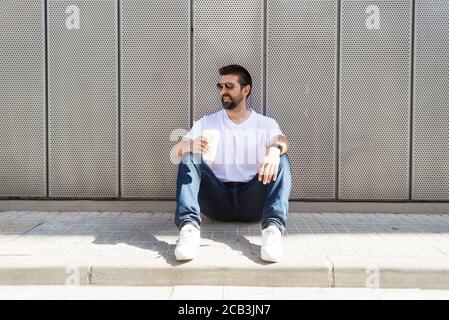 Ragazzo bearded con gli occhiali da sole seduti a terra mentre tiene un togliere il caffè Foto Stock