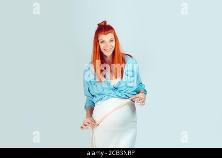 Felice giovane zenzero ragazza con incinta pancia che tiene il nastro di misurazione che si prende cura del peso e sorridendo a macchina fotografica isolata su blu. In buona forma con go Foto Stock