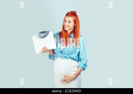 Affascinante donna rossa che si aspetta il bambino e toccando la pancia delicatamente in piedi con scale di peso e sorridendo alla fotocamera su blu. Dieta e cura del peso durante Foto Stock