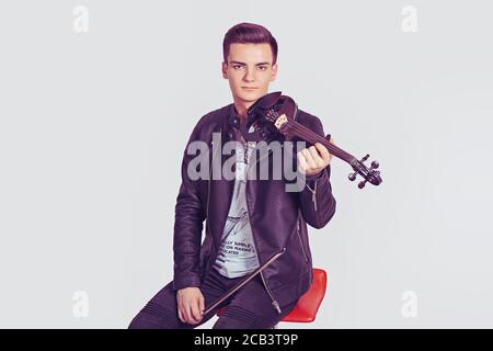 Giovane uomo in posa reggendo violino moderno e fiddle-stick sorridente seduto su una sedia rossa isolato su sfondo bianco parete. Modello con shot orizzontale in bla Foto Stock