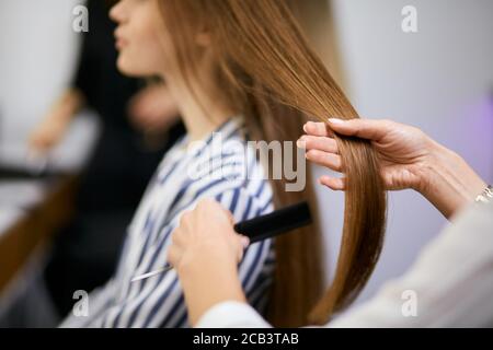 vista laterale sul parrucchiere corto tenendo e pettinando i capelli della giovane donna nel salone di bellezza, la giovane donna ha i capelli lunghi. Bellezza, concetto di capelli Foto Stock