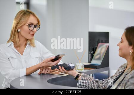 bella donna con capelli biondi corti pagare per le procedure di bellezza in salone professionale, sorridente personale cordiale alla reception Foto Stock