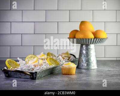 Mini cupcake al limone con crema bianca e gialla seduta in un vassoio metallico guarnito con fettine artificiali di limone e. un rustico in metallo rialzato di Foto Stock