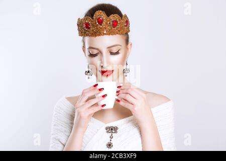 Gustando la sua tazza di tè del caffè. Sorridente bellezza regina principessa sposa di essere bella donna con tazza bianca di bevanda calda in mano isolato su bianco gra Foto Stock