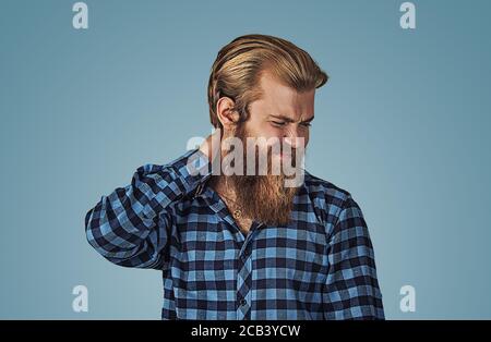 Ho bisogno di un massaggio. Uomo frustrato che tiene la mano sul collo, avendo dolore sulla schiena. Hipster maschio con barba in camicia a scacchi blu plaid isolato sopra Foto Stock