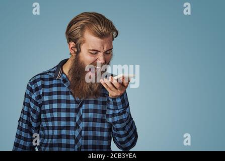 Ritratto giovane arrabbiato urlando sul telefono cellulare. Hipster maschio con barba in camicia a scacchi blu plaid isolato su sfondo blu studio. Negativ Foto Stock