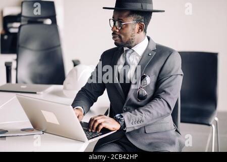Uomo d'affari sicuro africano in abito , occhiali e cappello dandy lavorando su laptop, analizzando gli ultimi rapporti da borsa. Foto Stock