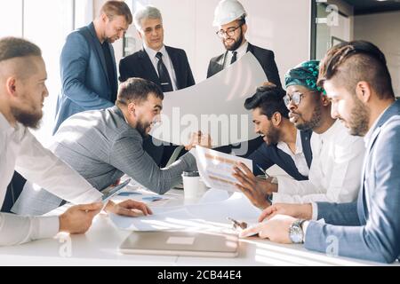 Gruppo multirazziale di costruttori, costruttori, genieri e architetti discutendo blueprint presso l'ufficio. Tutti gli uomini sono vestiti in abiti aziendali, e bianco Foto Stock