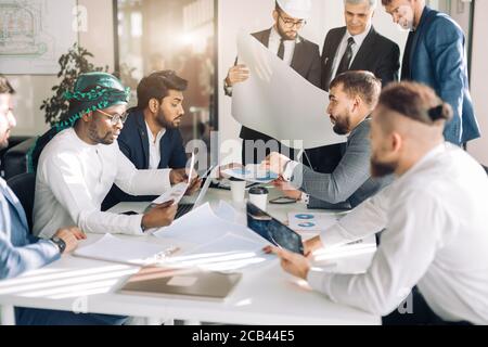 Focalizzato indiana, araba e caucasico ingegneri, con uno di loro indossa hardhat, discutendo la costruzione di grafici e progetti in costruzione compa Foto Stock