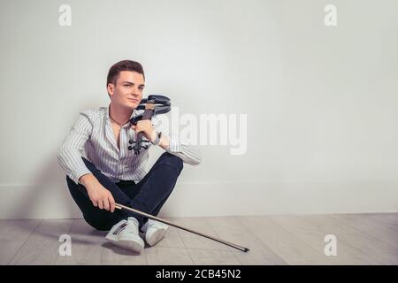 Bell'uomo giovane in camicia e denim che tiene il violino e. fiddle mentre si siede su sfondo bianco della parete e sognando di giorno Foto Stock